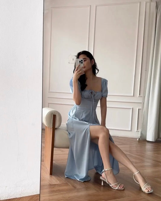 一位女士穿著來自 the.lookbook.select 的復古藍色 Adella 連身裙，坐在凳子上，在光線充足的房間裡對著鏡子擺姿勢自拍。
