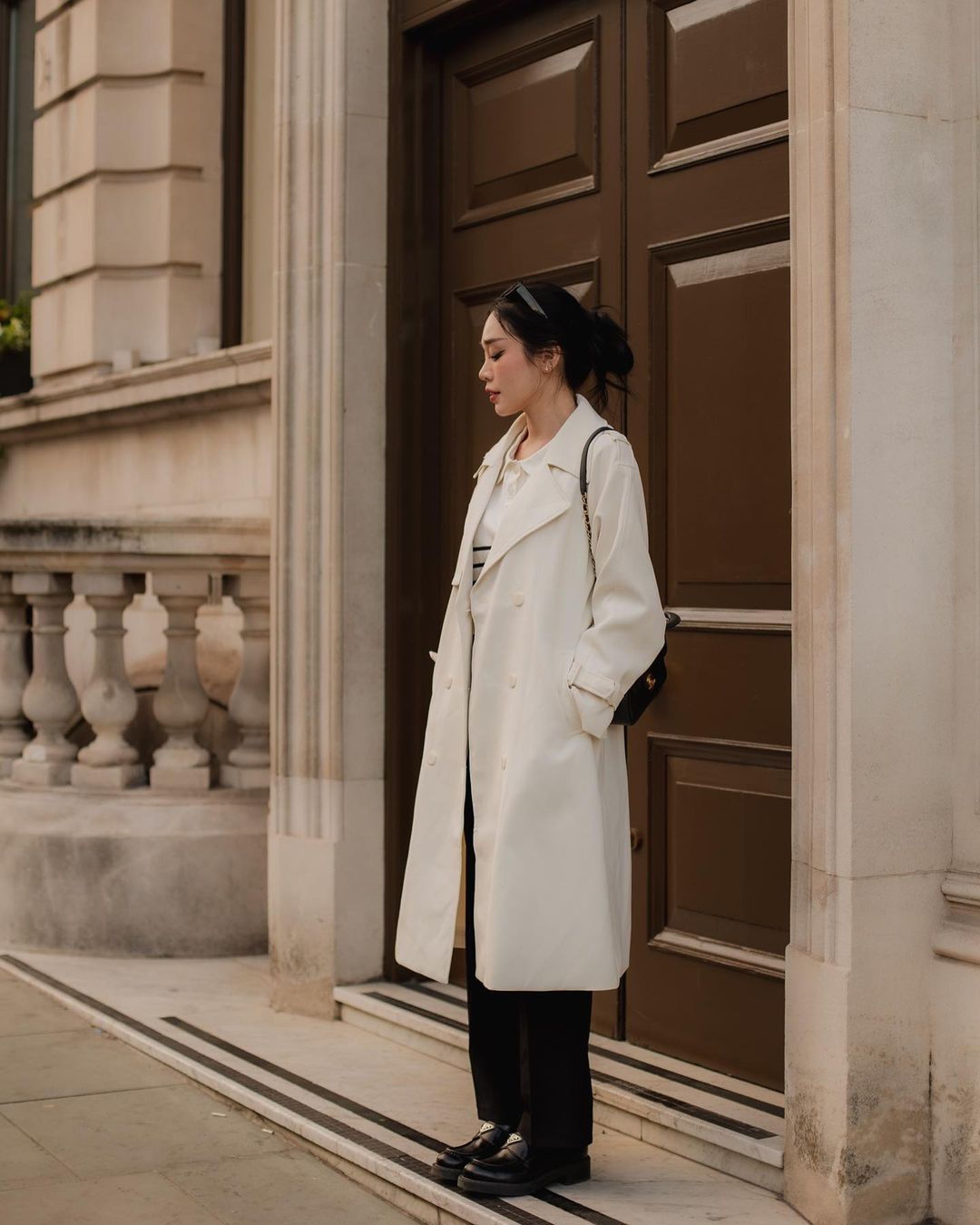 一位穿著 .lookbook.select 的白色經典 To-Go 風衣和黑色褲子的女士站在一扇棕色大門旁。