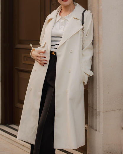 一位女士穿著 the.lookbook.select 的白色經典 To-Go 風衣，搭配條紋上衣和黑色褲子，一隻手放在腰上擺出姿勢。