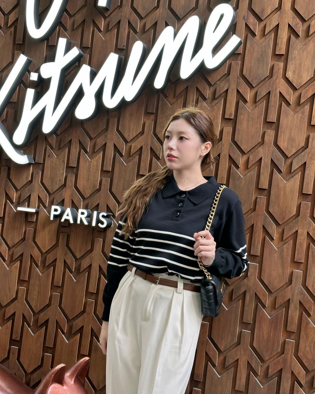 一位穿著 the.lookbook.select 舒適的 Danielle 毛衣的女士自信地站在寫有 Kitsume Paris 的標誌前，擁抱簡約的簡約設計和秋冬季節的疊搭技巧。