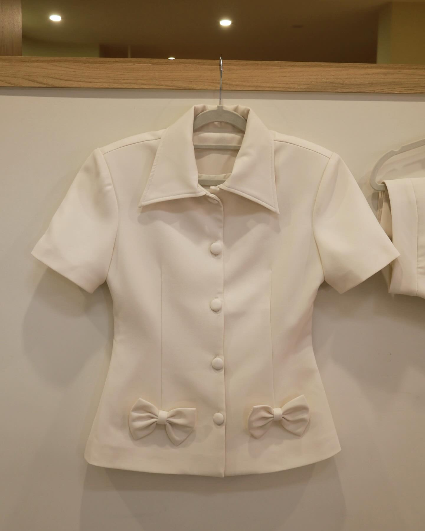 來自the.lookbook.select的Light Skirt Set，有一件乳白色的短袖上衣，前方有釦子和兩個裝飾性的腰部蝴蝶結，展示在衣架上。🖤新品優惠2件9折.