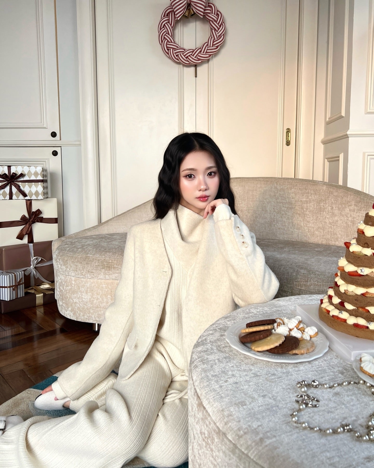 一位女士穿著 the.lookbook.select 的 Snow Wool Coat ❄️ 乳白色新品優惠，坐在米色沙發上的甜點旁，背景是包裝好的禮物和花圈。.