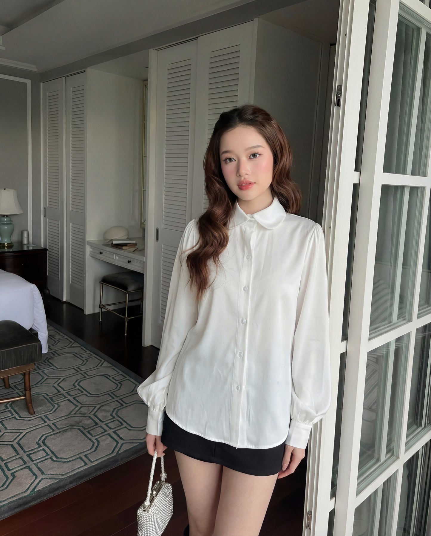 一位年輕女性穿著 The.lookbook.select Puff Sleeve Shirt 🖤新品優惠2件9折搭配黑色短褲，站在室內玻璃門附近，手上拿著質感十足的白色手提包。.