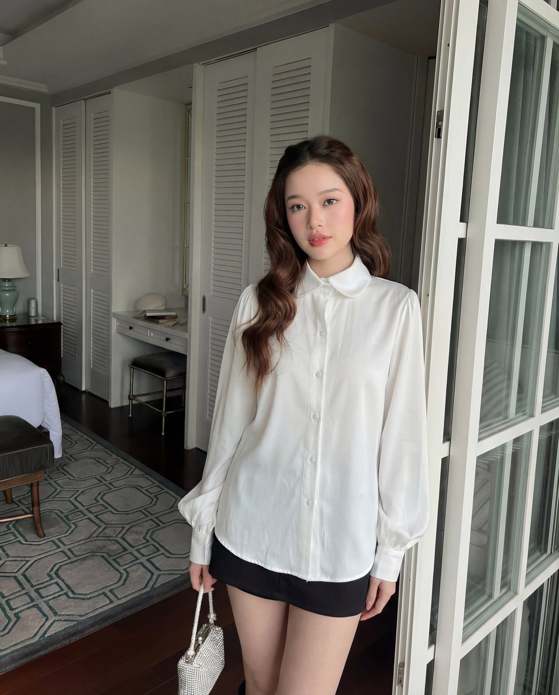 一位年輕女性穿著 The.lookbook.select Puff Sleeve Shirt 🖤新品優惠2件9折搭配黑色短褲，站在室內玻璃門附近，手上拿著質感十足的白色手提包。.