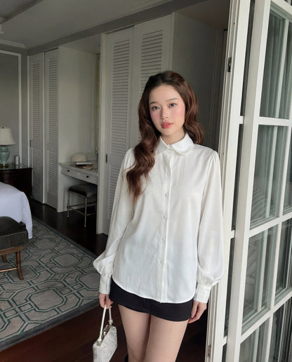 一位年輕女性穿著 The.lookbook.select Puff Sleeve Shirt 🖤新品優惠2件9折搭配黑色短褲，站在室內玻璃門附近，手上拿著質感十足的白色手提包。.