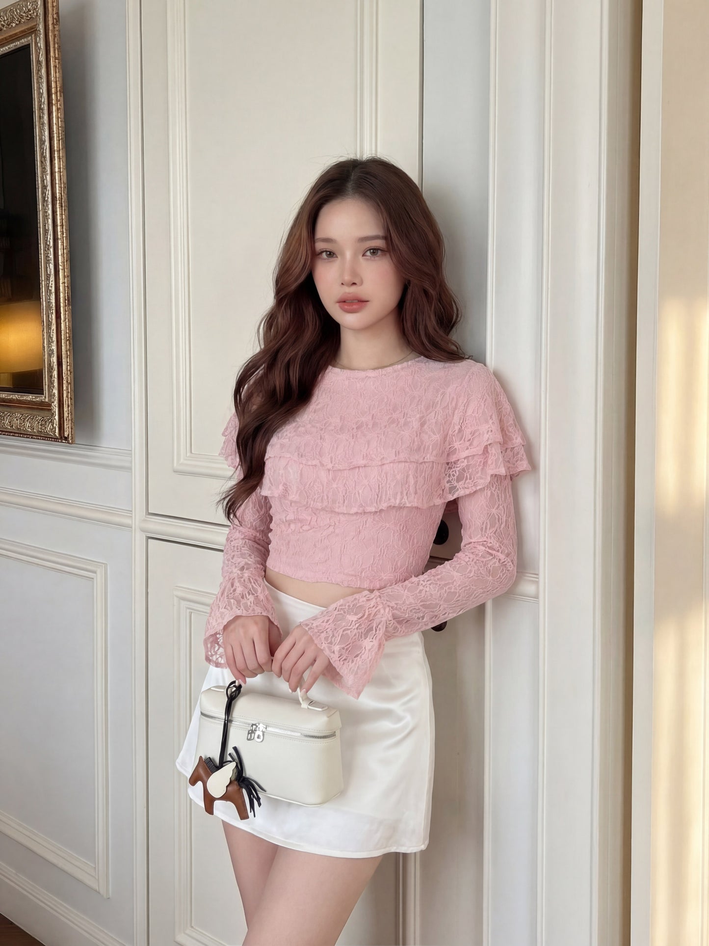一名身穿 the.lookbook.select 品牌粉紅色「Spring Muse」蕾絲上衣與白色裙子的女子站在室內，手拿白色手提包，望向鏡頭，那件浪漫的蕾絲上衣令她散發出一股優雅氣質。.