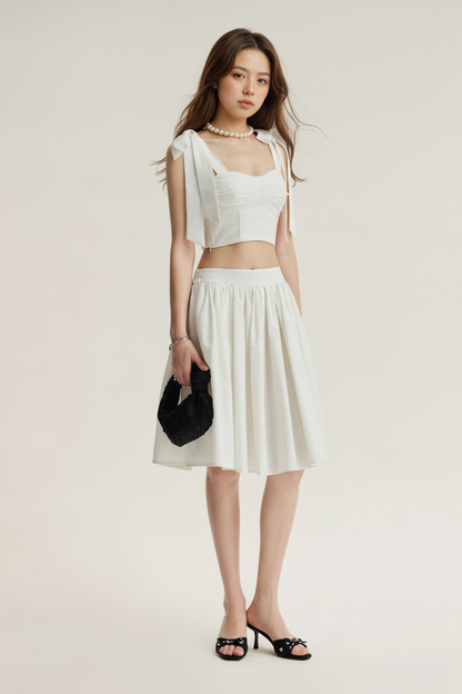 一位女士站在淺色背景下，身穿 the.lookbook.select 的 Jesse Midi Skirt、肩部系帶的白色 crop top、黑色涼鞋、珍珠項鍊，手拿黑色手拿包。.