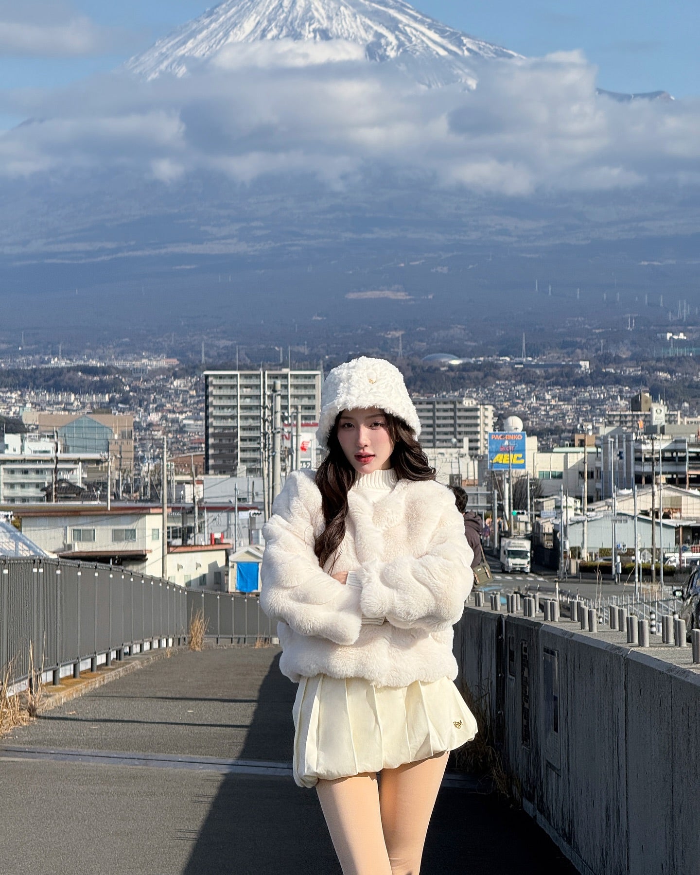 一位女士穿著 the.lookbook.select 的棉花糖皮草大衣 (附帽子)，站在人行道上，背景是建築物和白雪覆蓋的富士山。.