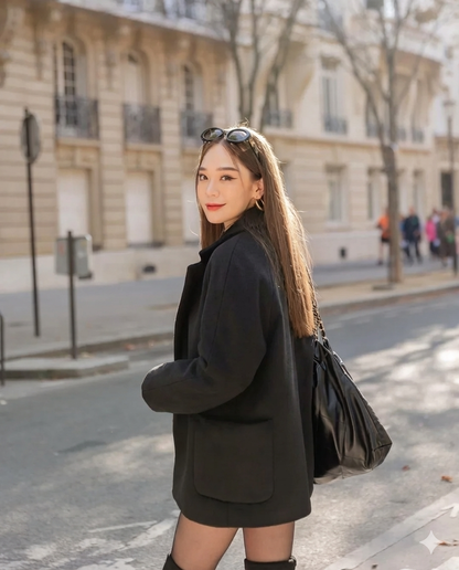 一位女士穿著 the.lookbook.select 的羊毛合身外套，挎著黑色包包，戴著太陽眼鏡，走在城市街道上，街道兩旁是淺色的建築物和光禿禿的樹木。.