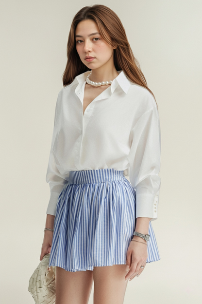 一位年輕女性穿著 the.lookbook.select 的 Lady Oversized Shirt，搭配藍白條紋短裙、珍珠項鍊和編織手袋。.