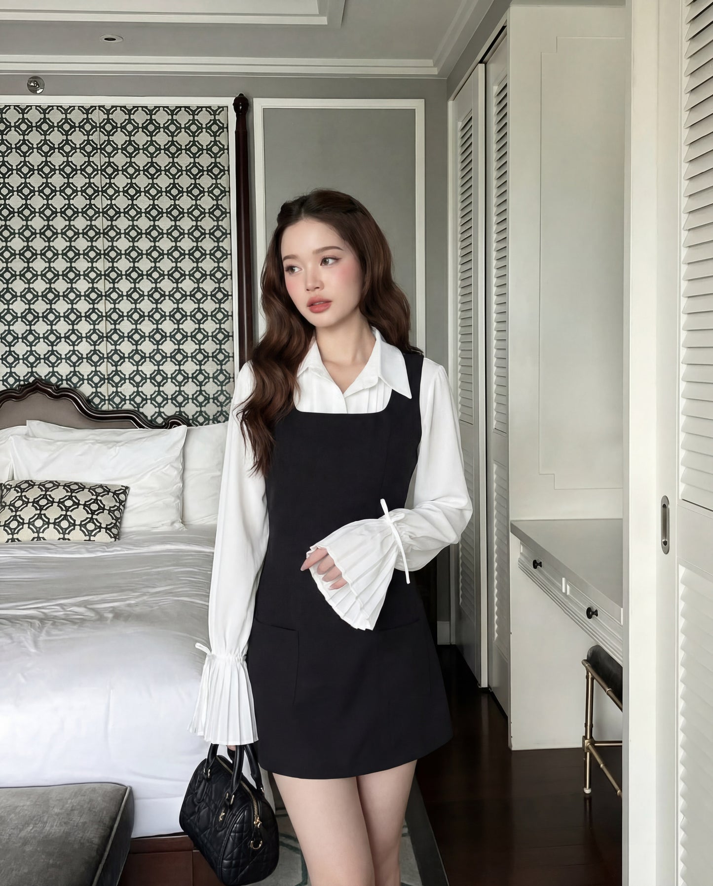 一位女士站在臥室中，身穿袖口打褶的白色上衣和 the.lookbook.select's Junior Sleeveless Dress，手拿黑色手提包。.