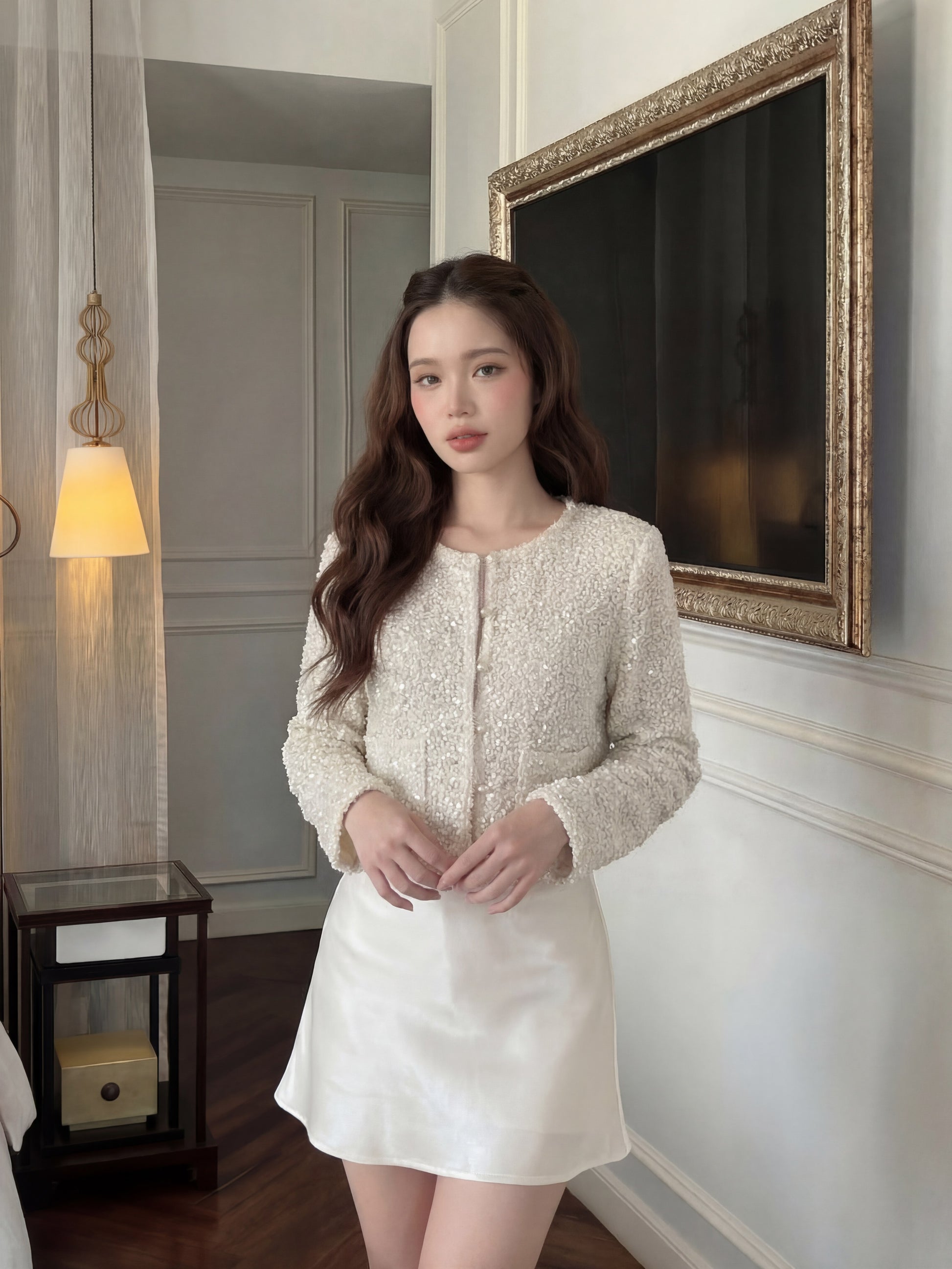 一名女子站在室內，身穿 the.lookbook.select 的「月光開襟衫」與白色裙子，她那波浪般的秀髮更添法式風情，身處這間佈置時尚的房間裡，室內擺放著一盞燈與裝裱好的藝術畫作。.