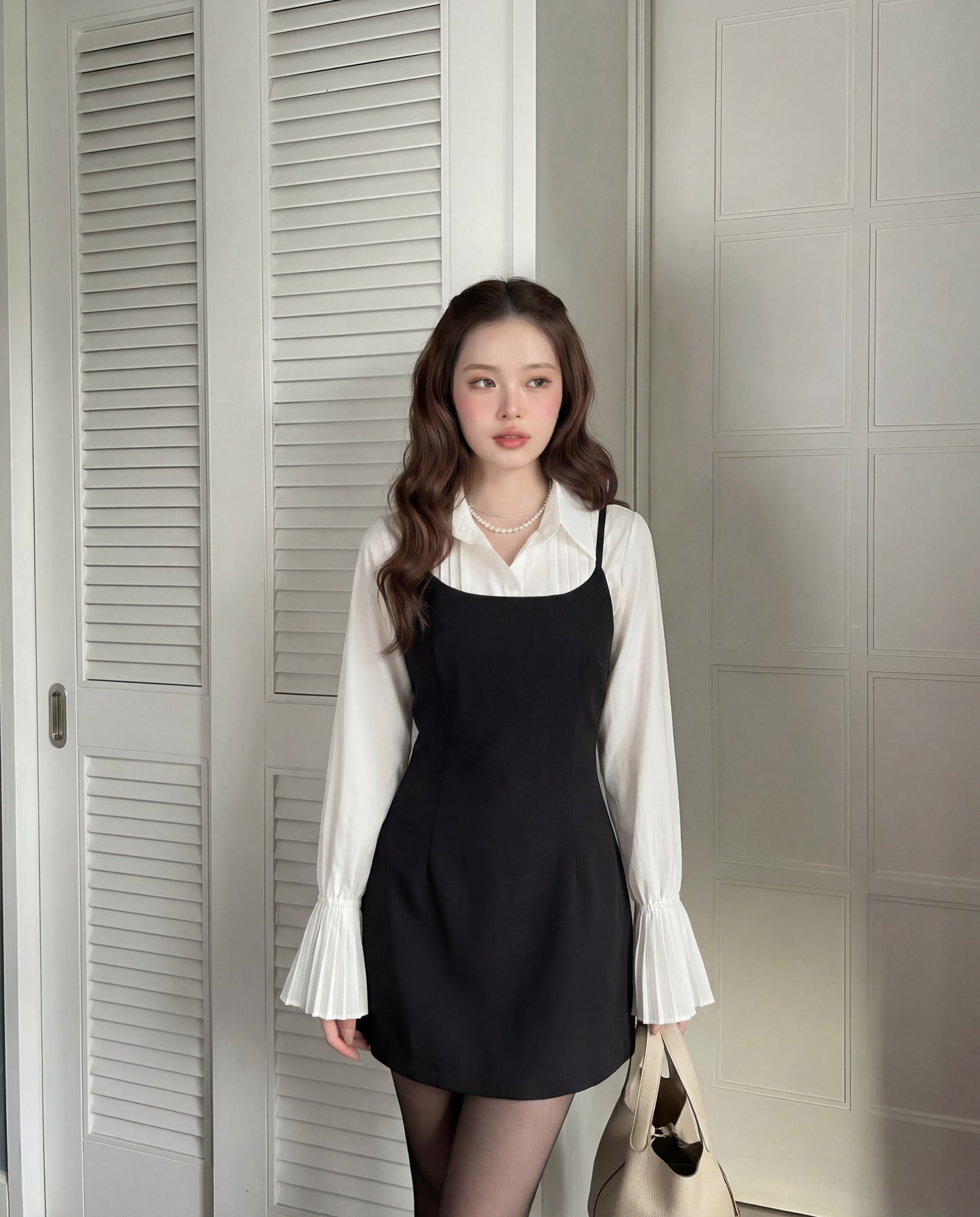 一位女士身穿 the.lookbook.select 的 Muse Cami 連身裙，外搭一件袖口有褶的白色上衣，搭配黑色緊身褲和米色手提包，站在室內白色嵌板門前擺姿勢。.