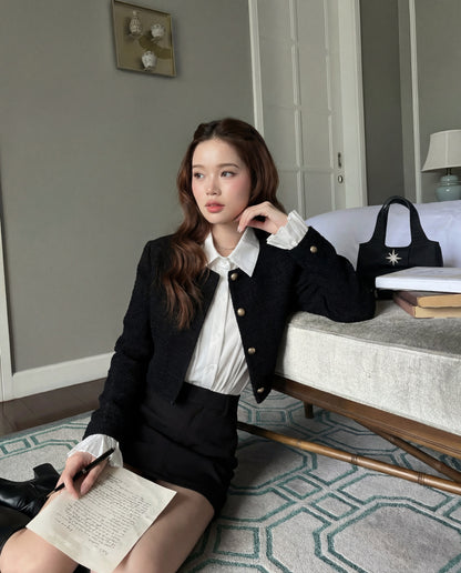 一位身穿 the.lookbook.select 黑色 Sine Tweed Coat 和白色上衣的女士坐在長椅旁的地板上，手上拿著一張手寫的紙張，長椅上放著書本和一個黑色袋子。.