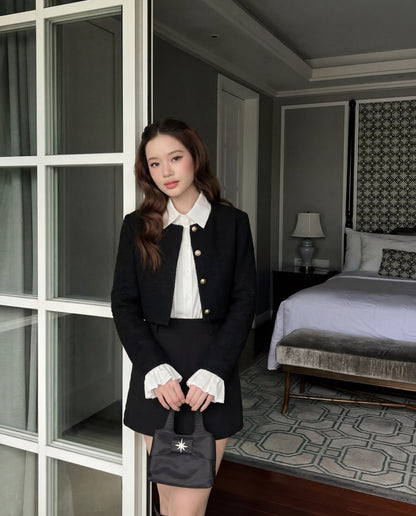 一位女士站在室內，身穿 the.lookbook.select 的 Sine Tweed Coat，外搭白色襯衫和黑色短裙，手拿黑色小包；背景可見整齊的床和台燈。.