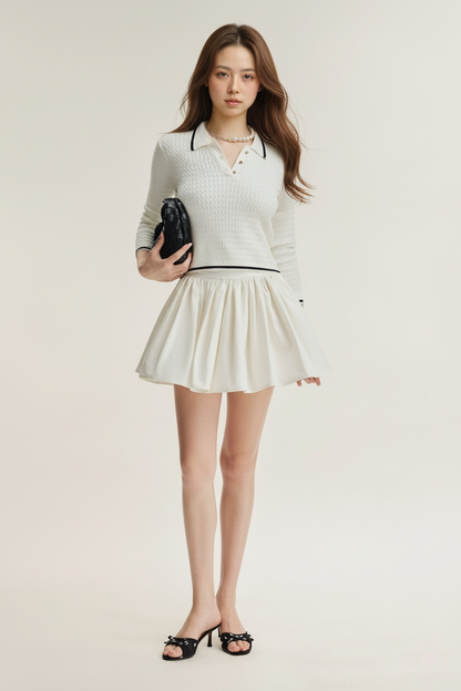 一位女士身穿 the.lookbook.select 的 Pierre 針織上衣，搭配白色喇叭裙和黑色高跟鞋，手拿黑色手拿包，站在素色背景下。.