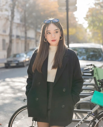 一位長直髮的女士站在戶外停放的腳踏車旁，身穿 the.lookbook.select 的羊毛合身外套，外搭白色上衣，配戴太陽眼鏡和金色環狀耳環，她的黑色外套在陽光普照的街道上更顯時尚。.