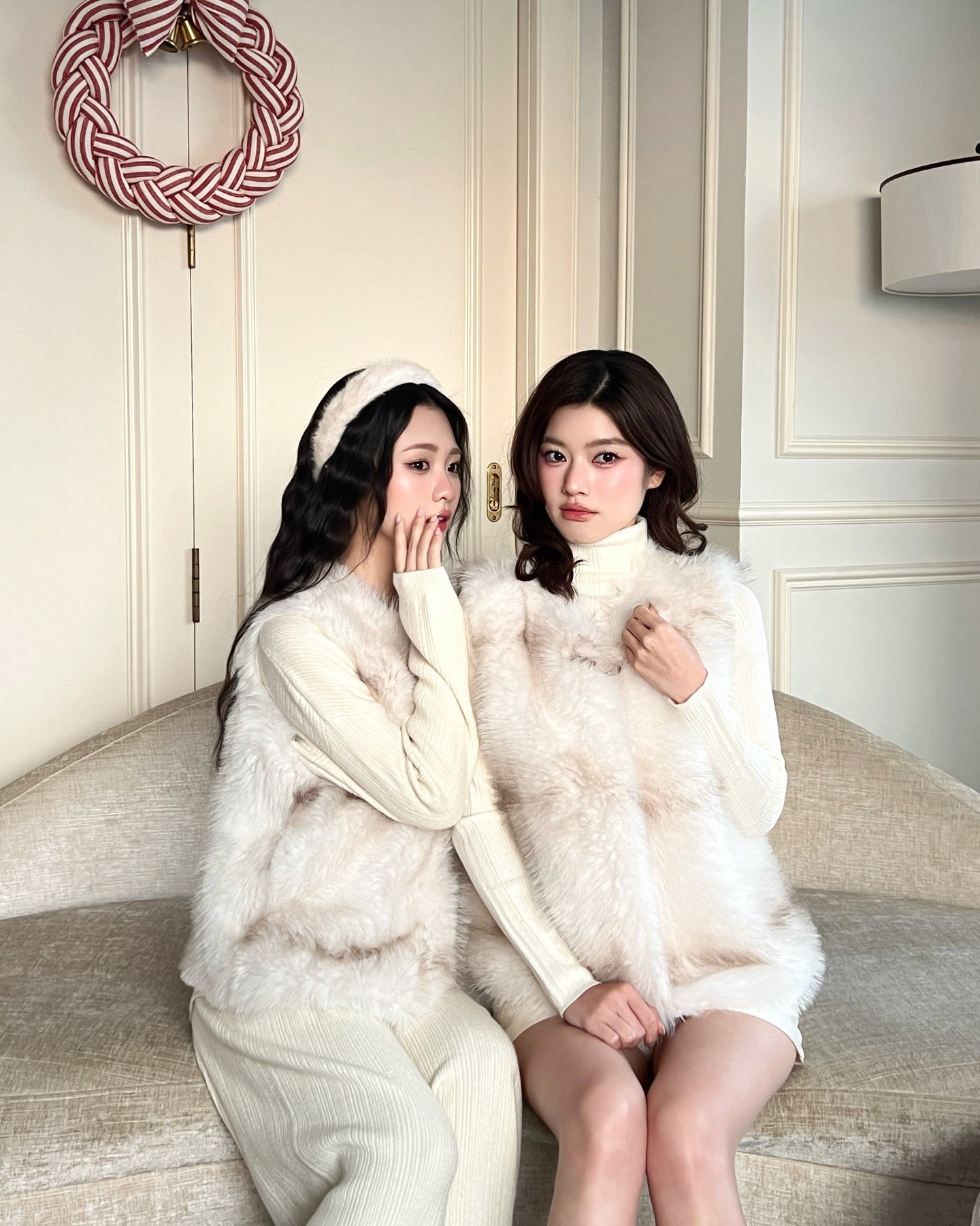 兩位穿著乳白色衣服和 La Neige Fur Vest ❄️ 新品優惠 by.lookbook.select 的女士坐在米色沙發上，房間內光線柔和，牆壁是白色嵌板，牆上掛著紅白相間的花圈。.