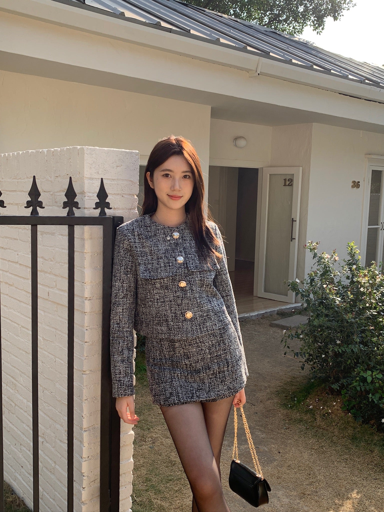 一位身穿 the.lookbook.select 的「This Girl Tweed Set」的人站在一棟白色房子外，倚著帶有黑色柵欄的白色磚牆，手裡拿著一個帶金鍊的黑色錢包。.