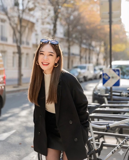 在陽光明媚的城市街道上，一位棕色長髮的女士身穿 the.lookbook.select 的羊毛合身外套，外搭白色上衣，頭戴太陽眼鏡，微笑地站在停放的腳踏車旁。.