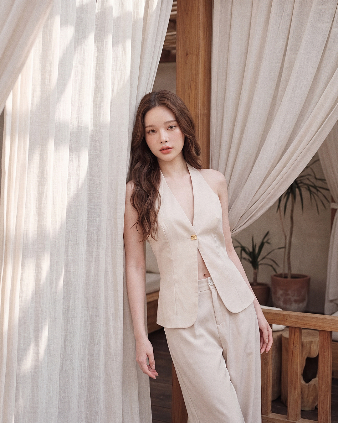 一名身穿由 the.lookbook.select 推出的「依雲度假套裝」的女子，倚靠著一扇掛著白色窗簾的木框窗，背景中可見盆栽植物。.