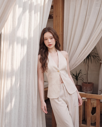 一名身穿由 the.lookbook.select 推出的「依雲度假套裝」的女子，倚靠著一扇掛著白色窗簾的木框窗，背景中可見盆栽植物。.