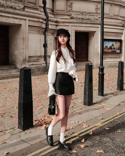 一位女士穿著 the.lookbook.select 的 Grace Mini Skirt 🖤新品優惠至 12 月 5 日，站在城市人行道的秋葉中，搭配白色上衣、黑色帽子和黑色手提包。.