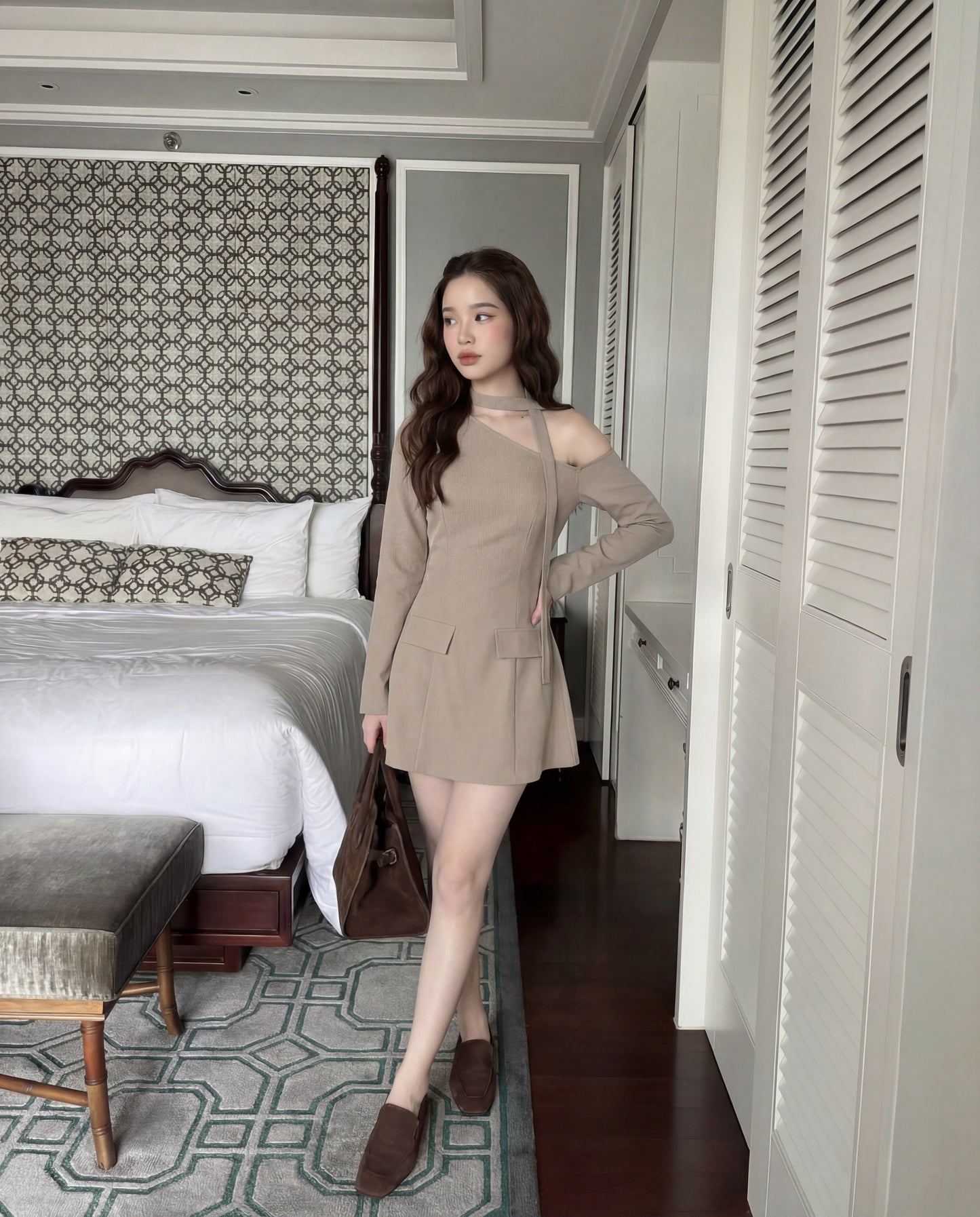 一位身穿 the.lookbook.select 單肩西裝連衣裙的女士站在現代化的臥室中，手持棕色手提包，床、長椅和花紋地毯清晰可見。.