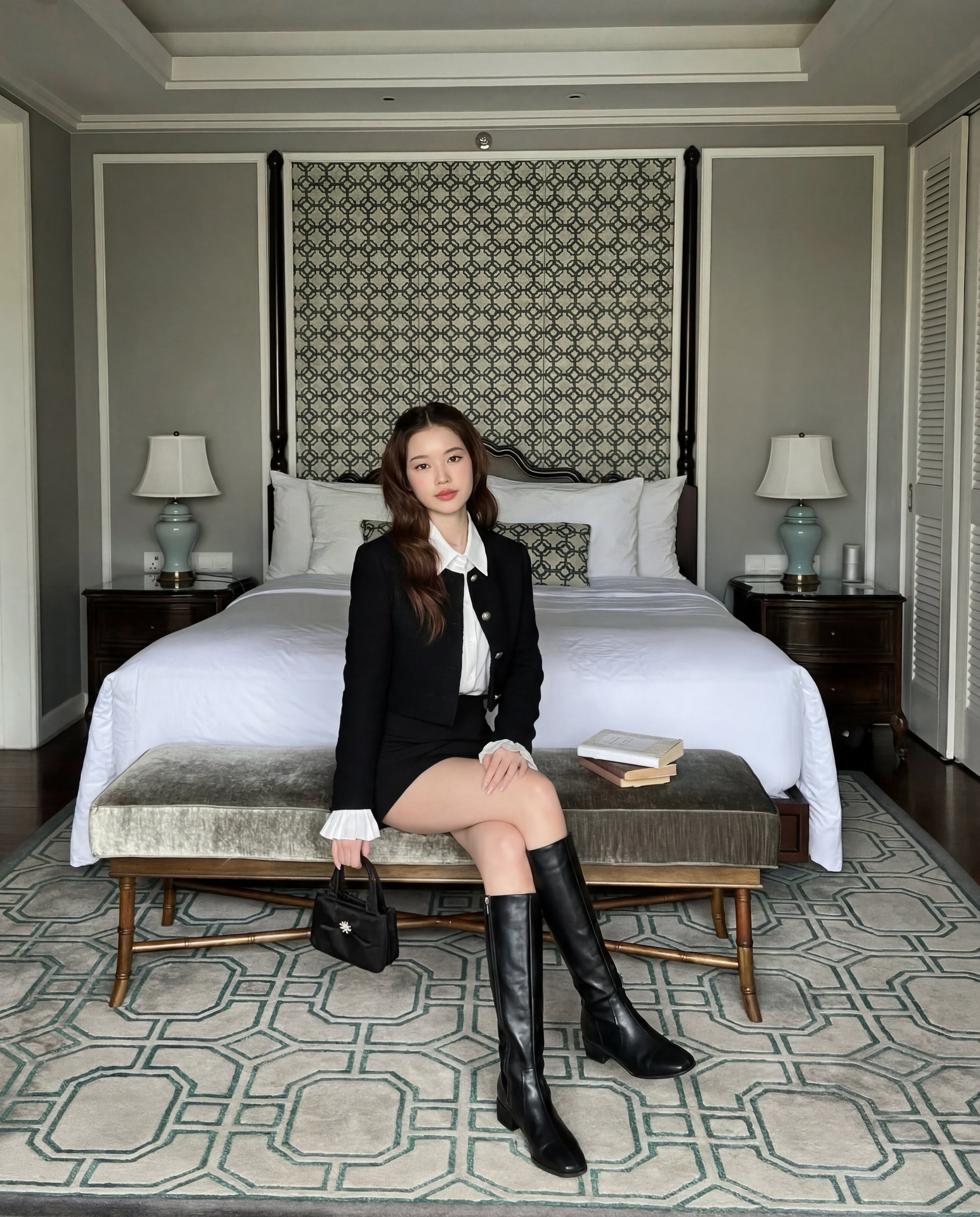 一位身穿 the.lookbook.select 黑色 Sine Tweed 大衣、白色襯衫和及膝長靴的女士，坐在裝潢現代、整齊的臥室床腳旁的長椅上，身旁放著兩本打開的書。.