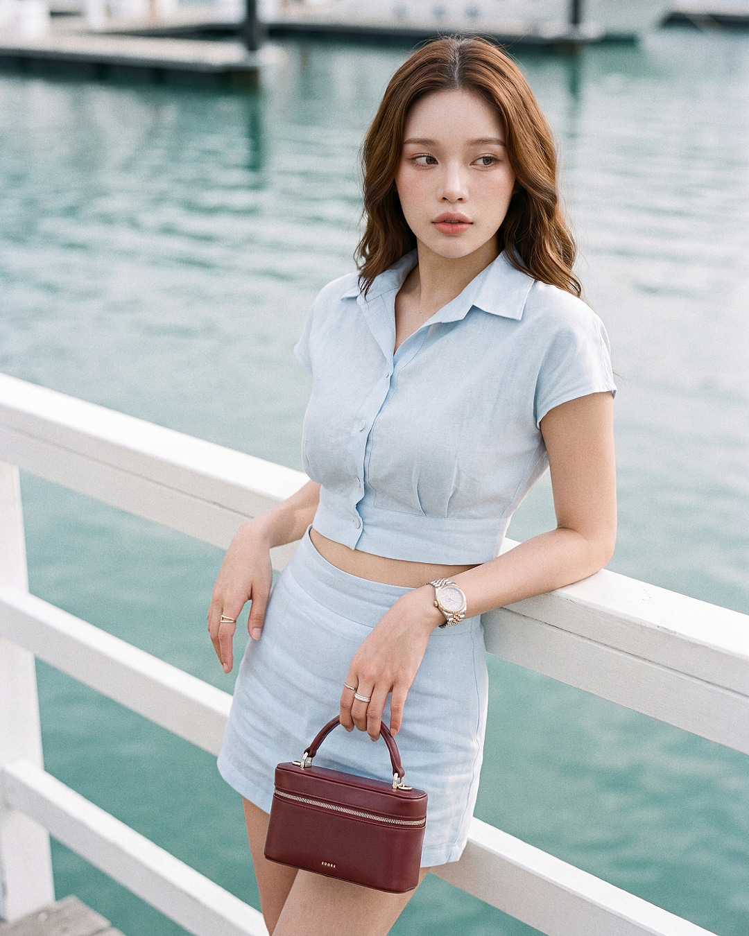 一位身穿.lookbook.select「Breeze 亞麻套裝」💌新品優惠：買兩件享九折——一套淺藍色短袖短版上衣與半身裙——的女子，站在水邊的白色欄杆旁，手拿一個小巧的酒紅色手提包。.