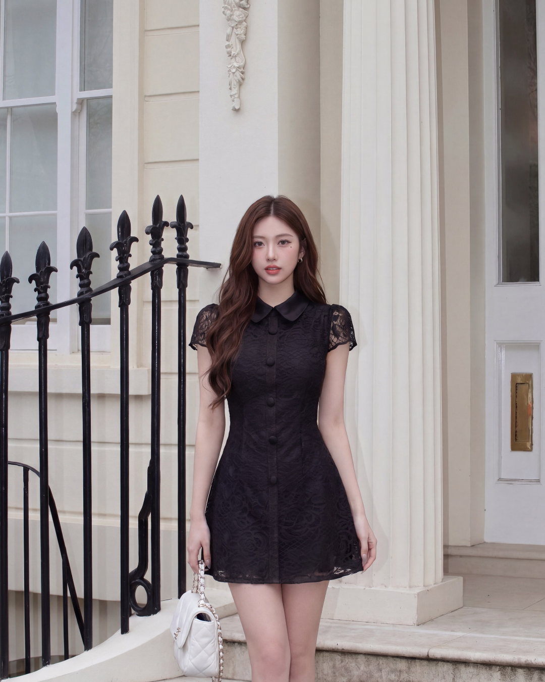 一位女士穿著 the.lookbook.select 的 Wellington 蕾絲連身裙，站在一棟有白色柱子和黑色鐵柵欄的建築物前，手上拿著一個白色手提包。.