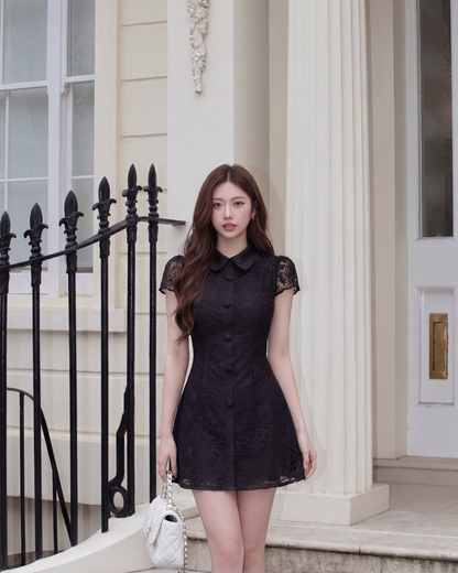 一位女士穿著 the.lookbook.select 的 Wellington 蕾絲連身裙，站在一棟有白色柱子和黑色鐵柵欄的建築物前，手上拿著一個白色手提包。.