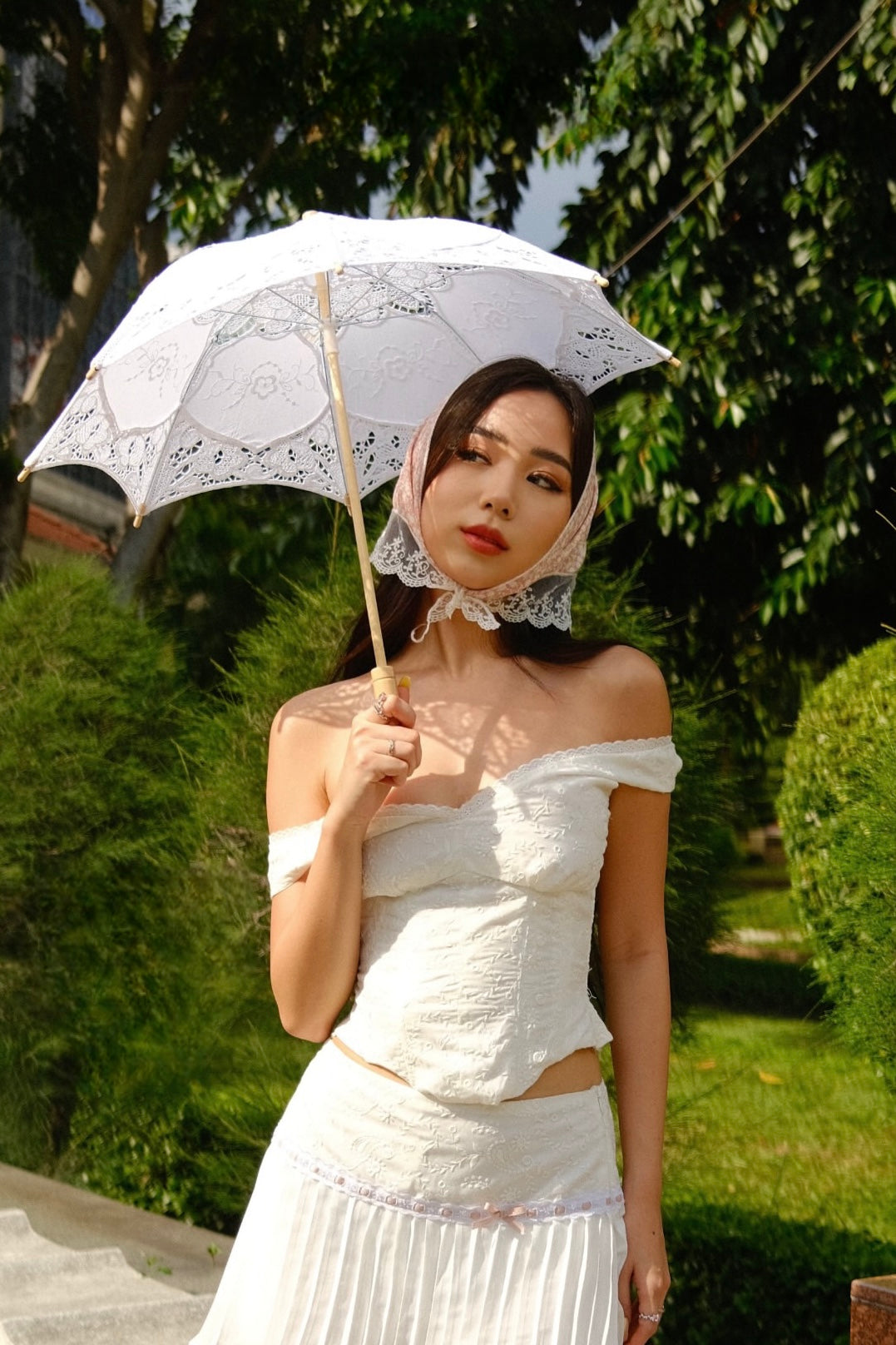 一位穿著 the.lookbook.select 的 Amour Set 的女士，身穿飾有少年小花的白色露肩連身裙，頭戴蕾絲頭巾，手持蕾絲遮陽傘站在戶外綠樹叢中。