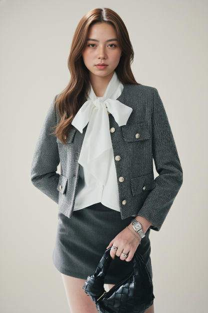 一位女士站在中性背景下，身穿 the.lookbook.select 的 Bow 寬鬆襯衫，搭配金色鈕扣的灰色外套和灰色短裙，手持黑色編織手袋。.