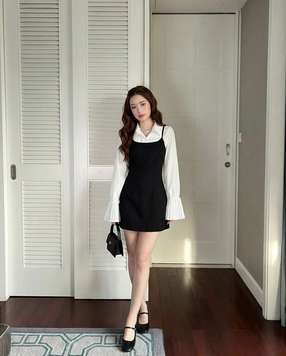 一位女士站在室內，身穿 The.lookbook.select 的喇叭袖襯衫，內搭黑色無袖連身裙，手拿黑色小手包，腳穿黑色皮鞋。.