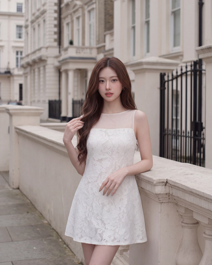 一位留著長棕髮的女子身穿由 the.lookbook.select 推出的「Elysee 蕾絲迷你洋裝」，佇立在兩旁盡是白色建築物的城市街道上，身後是石製欄杆。.