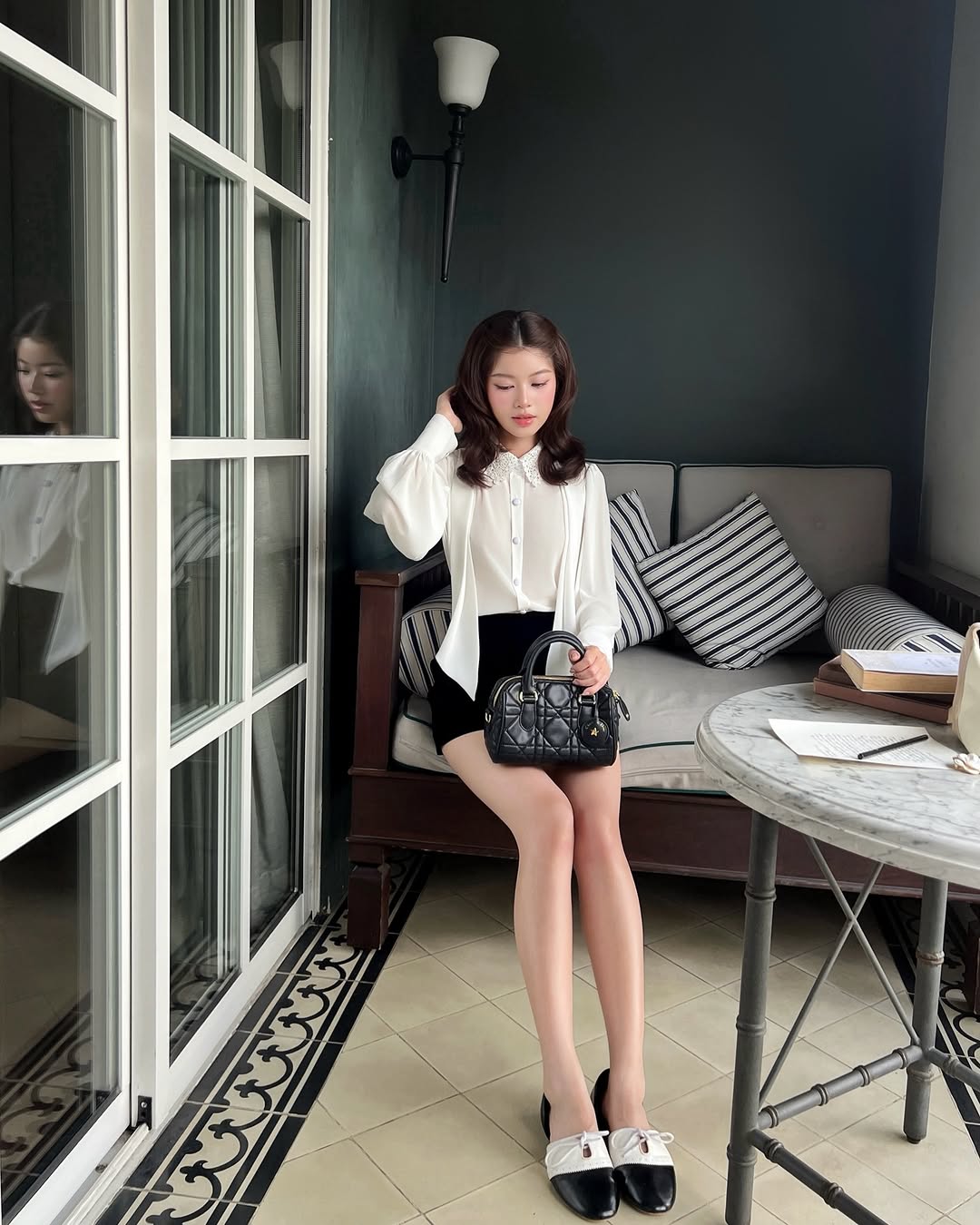 一位女士坐在桌子旁的軟墊長椅上，手拿黑色手提包。她穿著 the.lookbook.select's Heart White Shirt ❄️ 新品優惠搭配黑色短褲，身後有條紋枕頭，玻璃上可見她的倒影。.