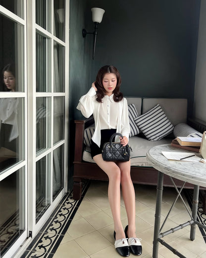 一位女士坐在桌子旁的軟墊長椅上，手拿黑色手提包。她穿著 the.lookbook.select's Heart White Shirt ❄️ 新品優惠搭配黑色短褲，身後有條紋枕頭，玻璃上可見她的倒影。.