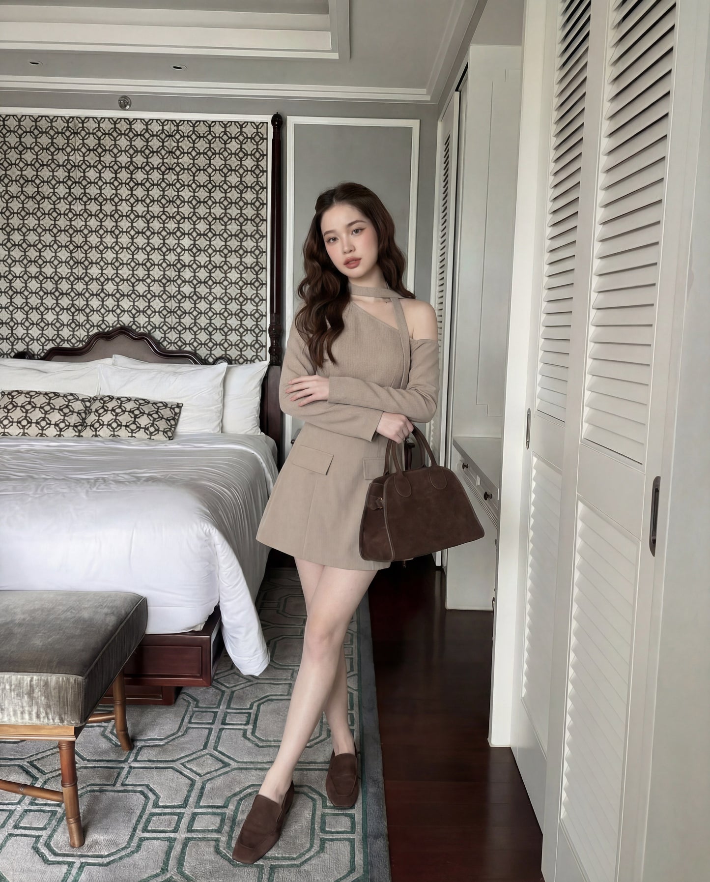 一位女士站在時尚的臥室中，身穿 the.lookbook.select 的單肩西裝連衣裙，搭配同色手袋和棕色鞋子，在床和衣櫃附近擺姿勢。.
