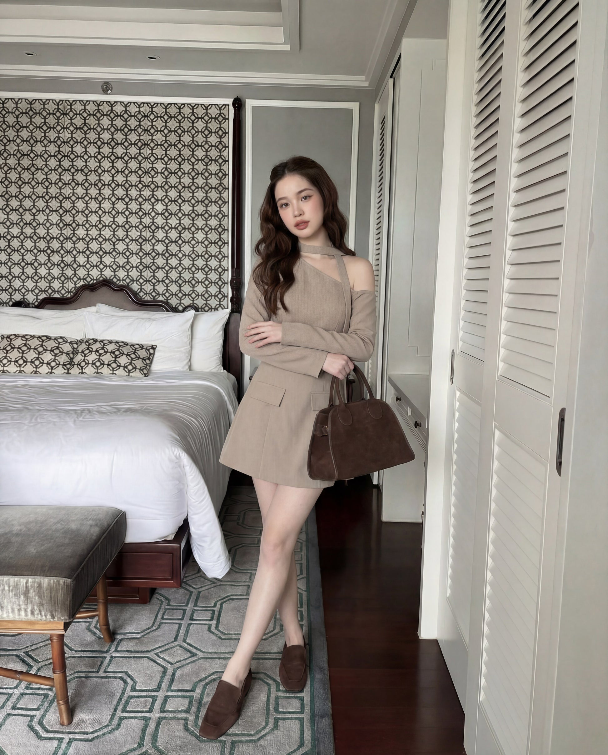 一位女士站在時尚的臥室中，身穿 the.lookbook.select 的單肩西裝連衣裙，搭配同色手袋和棕色鞋子，在床和衣櫃附近擺姿勢。.