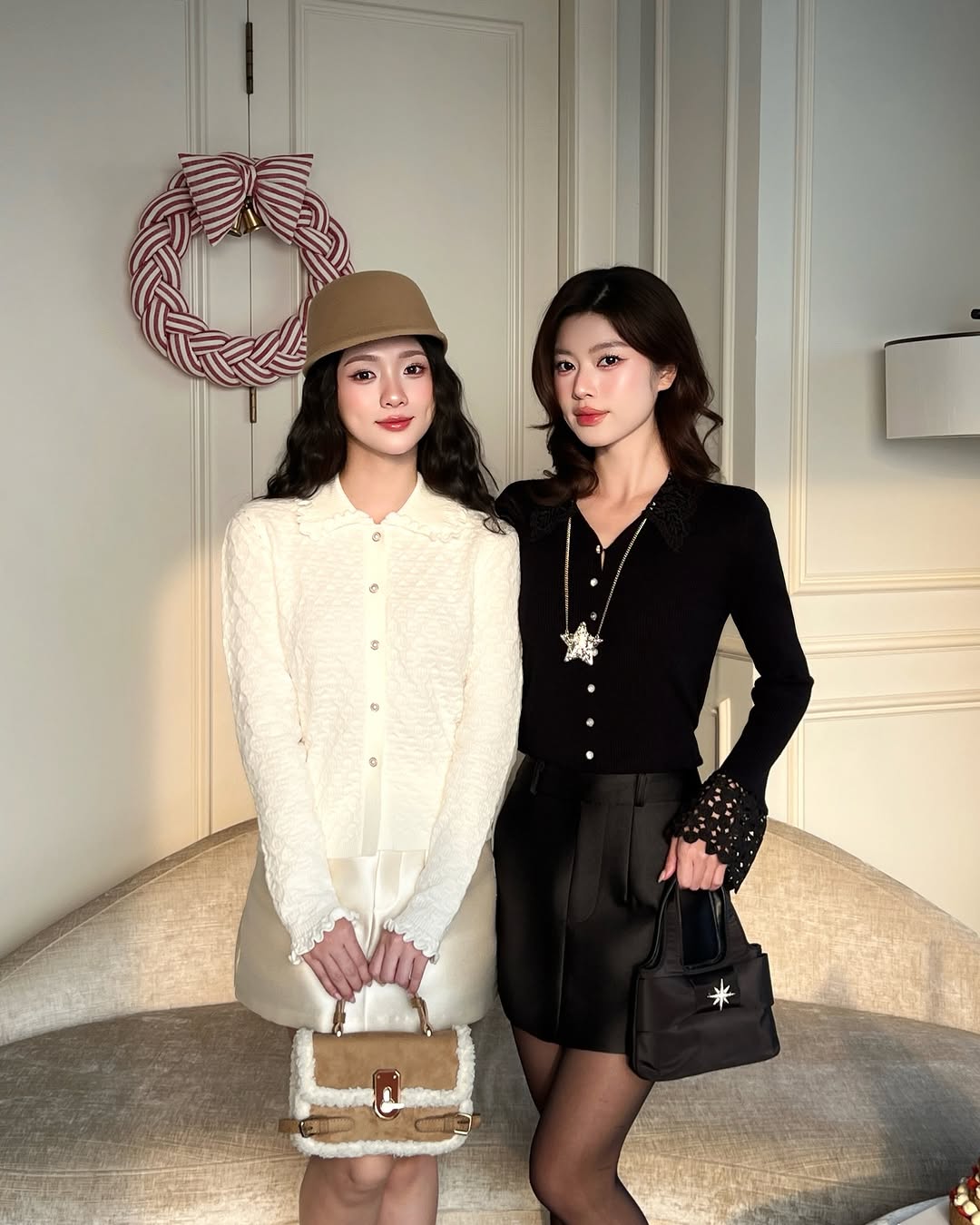 兩個女人在室內的米色沙發前擺姿勢。其中一位穿著 the.lookbook.select 的蕾絲針織上衣❄️，搭配一頂棕褐色帽子，另一位則是黑色合身針織上衣和短裙。他們身後的牆上掛著裝飾性的花圈。.