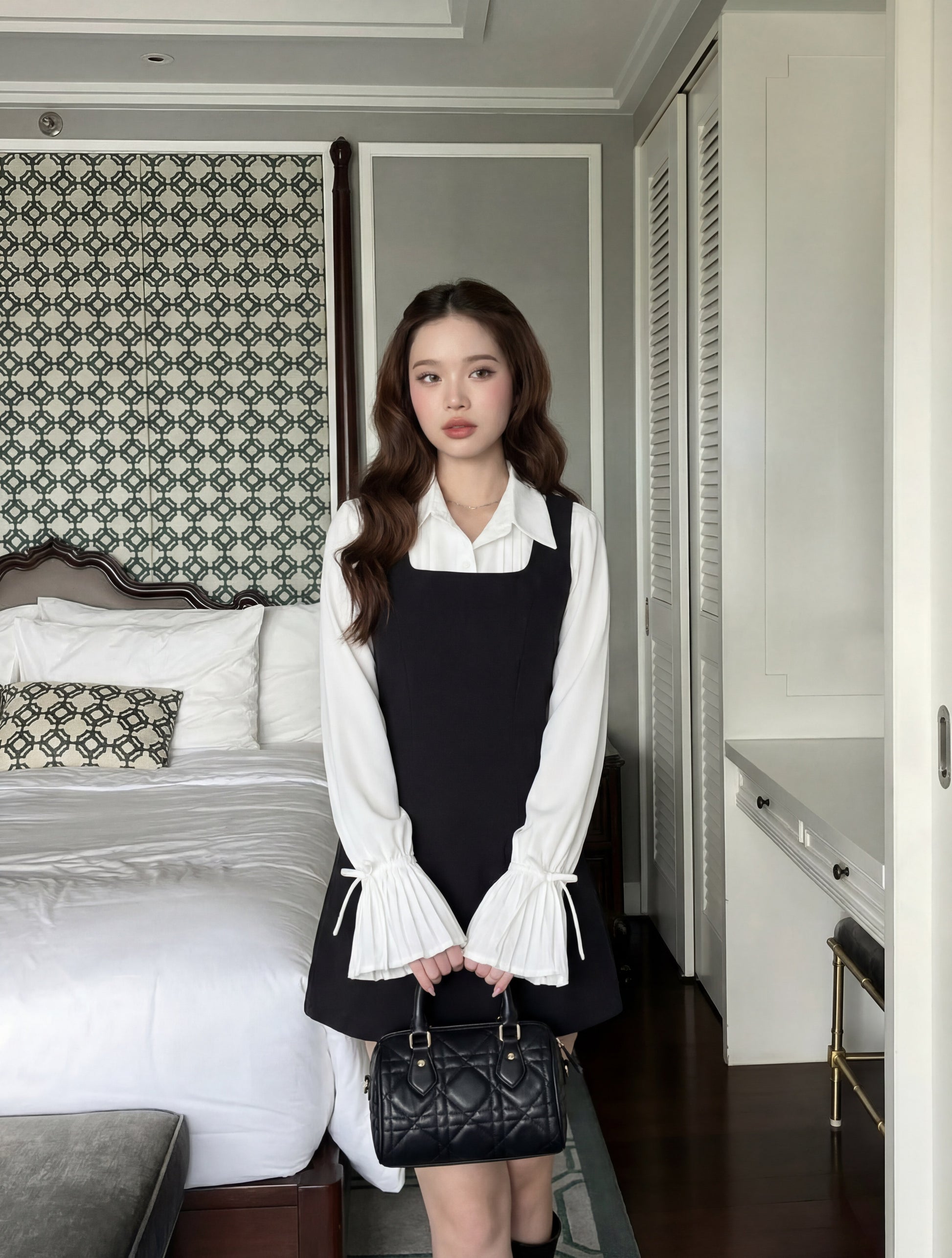 一位年輕的女性站在室內的床邊，身穿 the.lookbook.select 的 Junior Sleeveless Dress 黑色連衣裙，外面套著一件喇叭袖的白色上衣，手上拿著一個黑色手提包。.