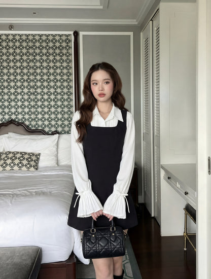 一位年輕的女性站在室內的床邊，身穿 the.lookbook.select 的 Junior Sleeveless Dress 黑色連衣裙，外面套著一件喇叭袖的白色上衣，手上拿著一個黑色手提包。.