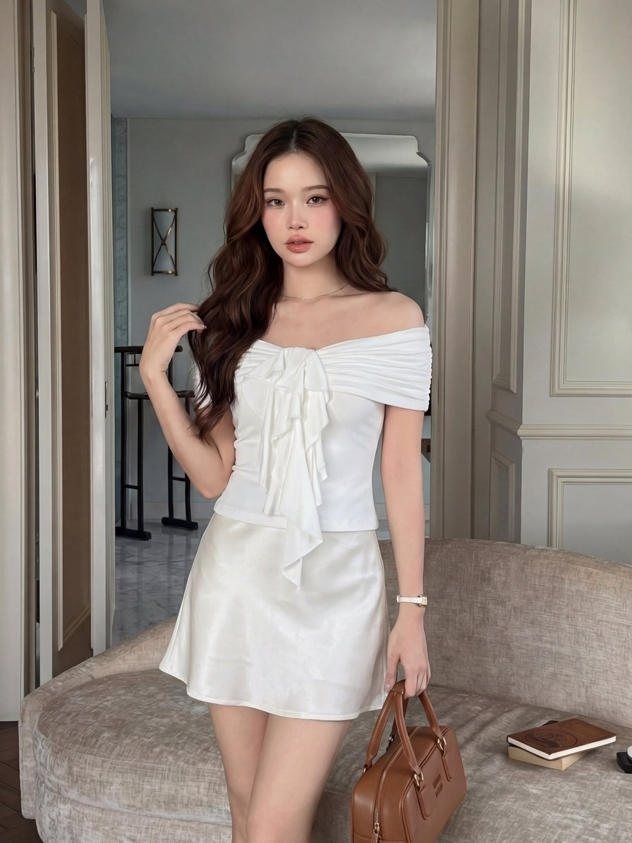 一位女士站在室內的米色沙發上，身穿 the.lookbook.select 的 Petal Bardot Top 搭配白色短裙，手持棕色手提包。.