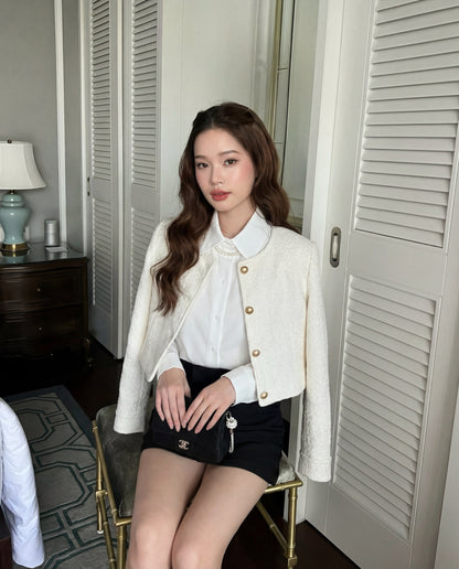 一位留著波浪長髮的女士坐在室內的椅子上，身穿白色上衣、the.lookbook.select 的 Sine Tweed Coat（金色鈕扣）和黑色短裙，手上拿著一個小皮包。.