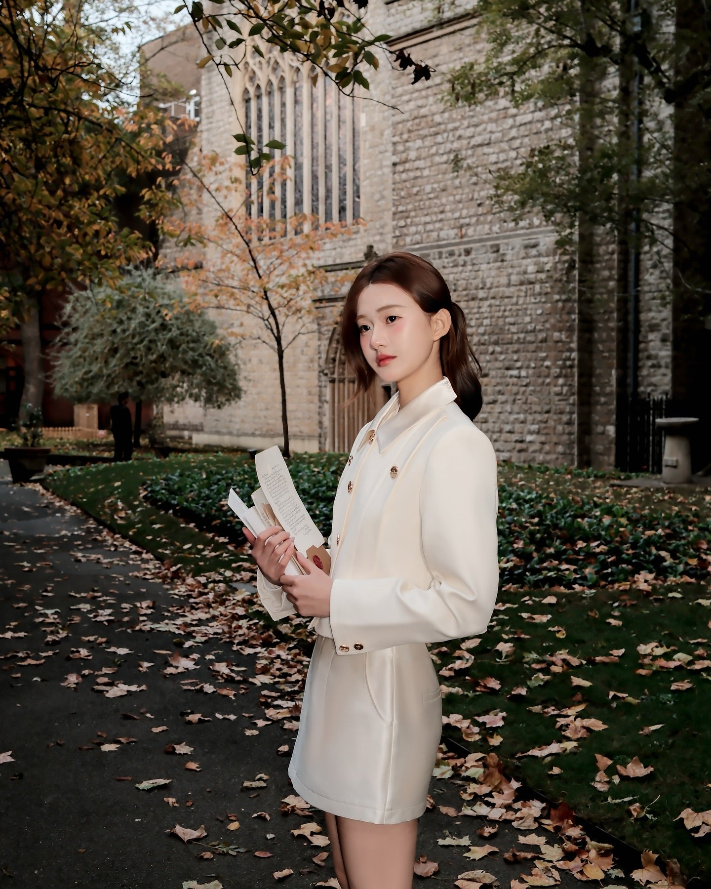 The.lookbook.select的Grace Little Jacket 🖤新品優惠至12月5日，一位女士站在戶外落葉鋪成的小路上，手持一本打開的書，背景是石頭建築和樹木。.