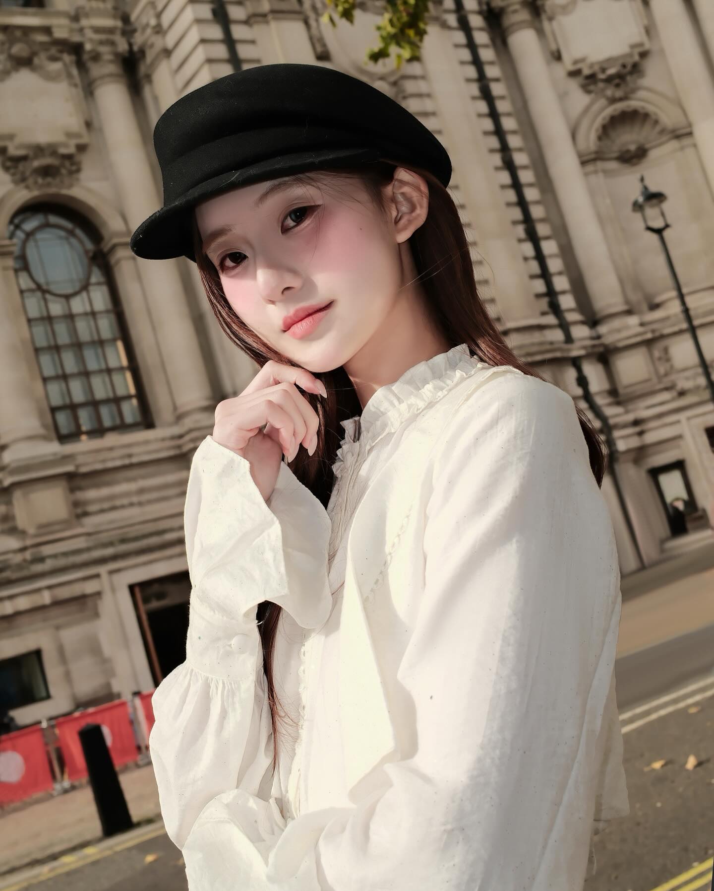 一位年輕女性穿著 Lookbook.select 的 Hazel White Shirt 🖤新品優惠至 12 月 5 日，戴著黑色帽子，在戶外的華麗石造建築中擺出造型，散發 法式氣質。.