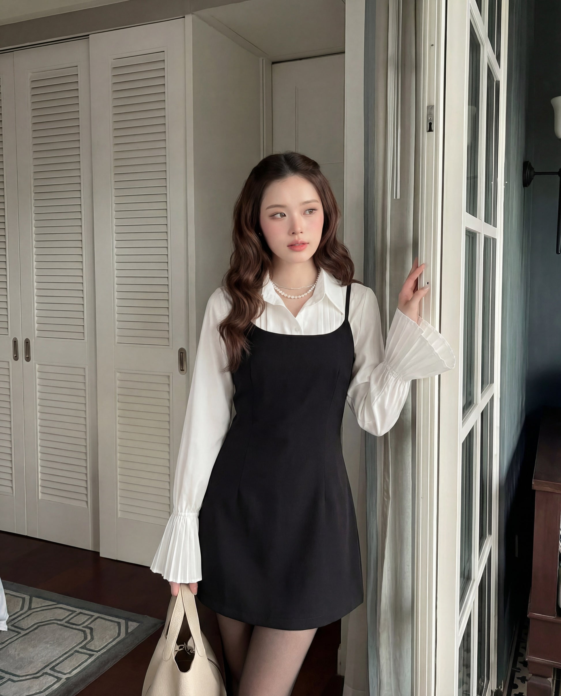 一位女士站在室內的玻璃門旁，在 the.lookbook.select 的 Muse Cami Dress 下穿著一件有褶袖的白色上衣，並拿著一個米色手提包。.