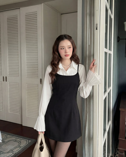 一位女士站在室內的玻璃門旁，在 the.lookbook.select 的 Muse Cami Dress 下穿著一件有褶袖的白色上衣，並拿著一個米色手提包。.