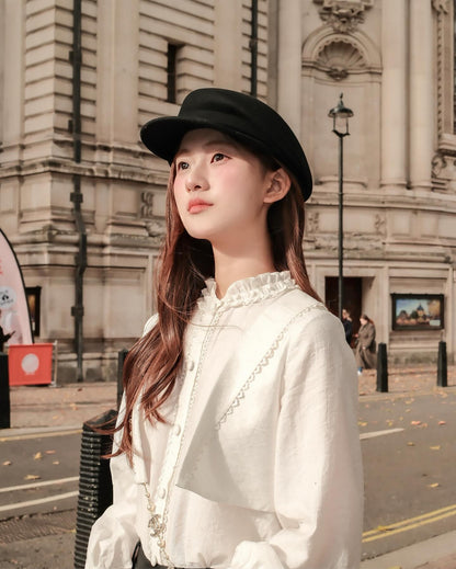 一位年輕女性穿著 the.lookbook.select 的 Hazel White Shirt 🖤，戴著黑色帽子，站在戶外一座宏偉的石造建築前，散發出毫不費力的法式氣質。.